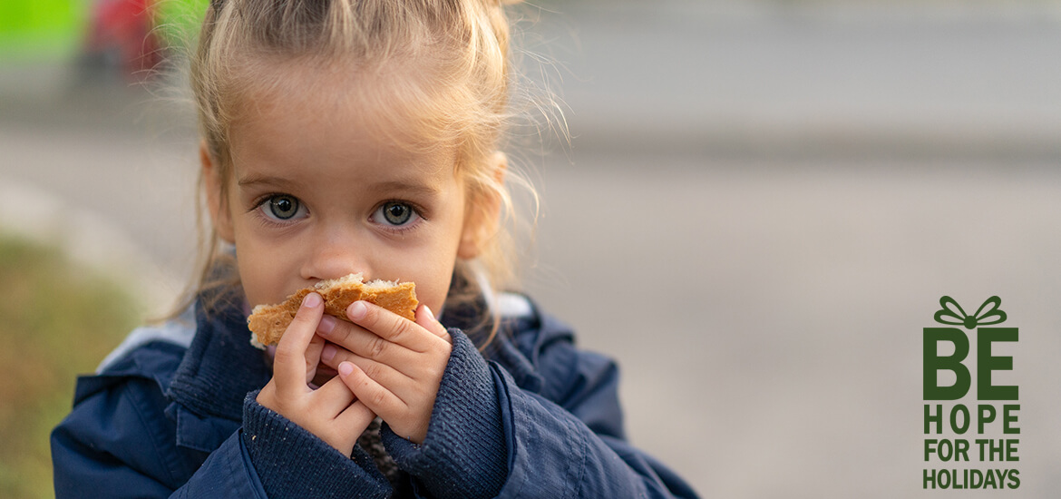 Young caucasian girl outside during winter, eating a piece of bread. Words say: Be Hope for the Holidays